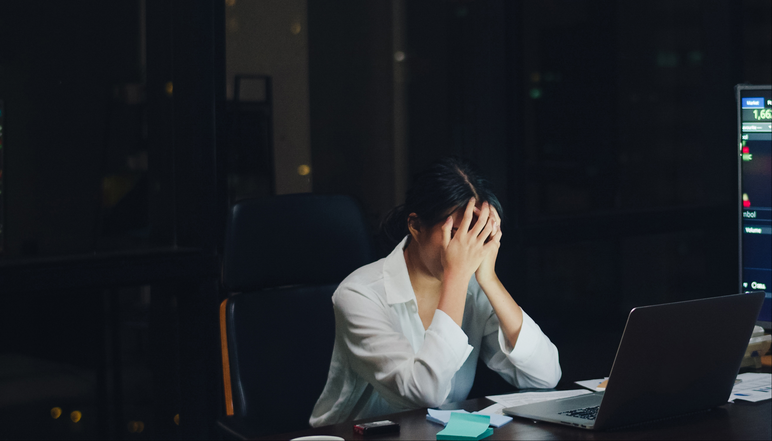 Mulher preocupada, com mãos no rosto, no escuro, no ambiente de trabalho.