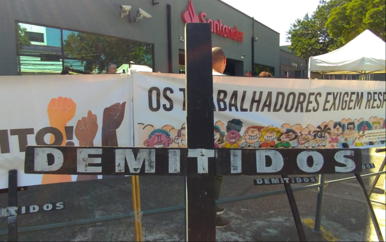 Imagem do protesto do Sindicato contra as demissões no Santander. A foto mostra uma cruz negra onde se lê "demitidos" e, atrás dela, faixas em frente à fachada do centro istrativo
