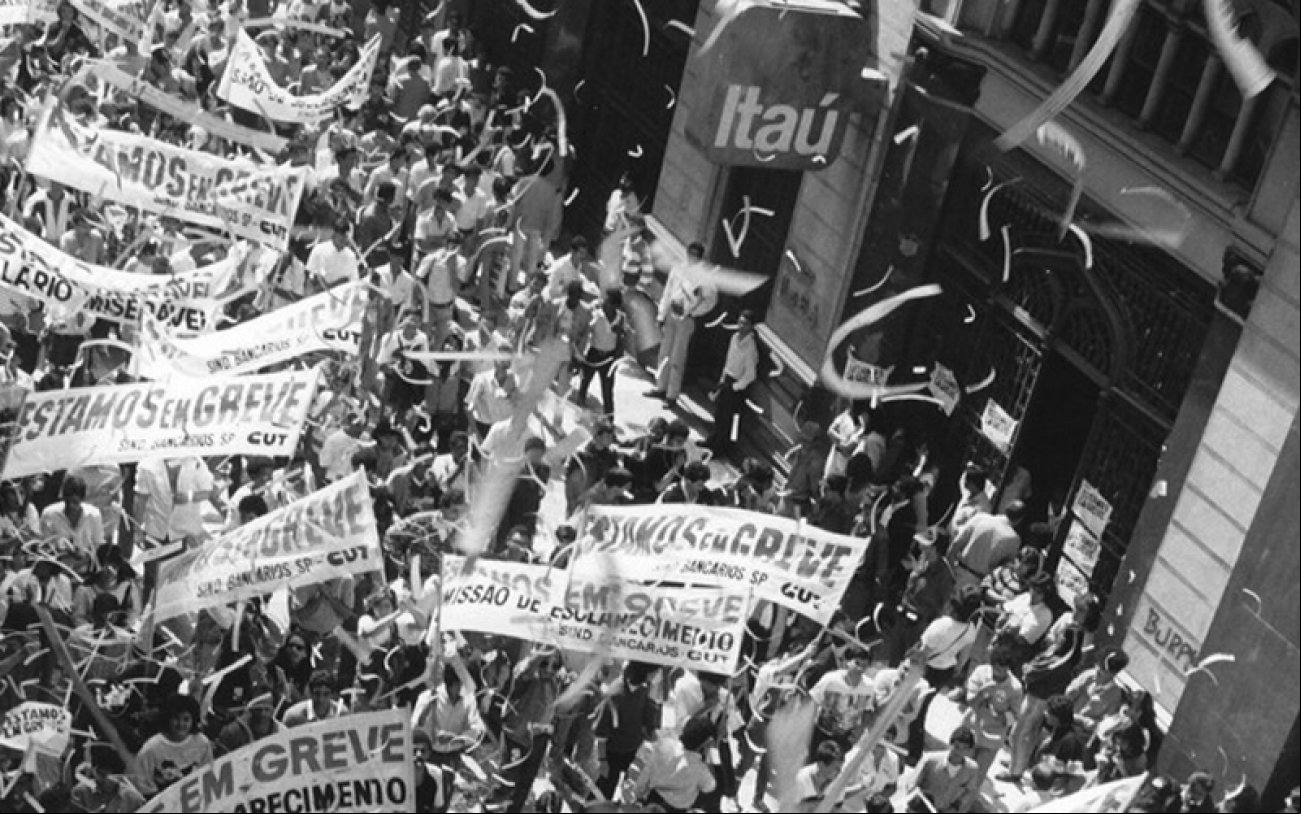 Bancários protestam durante greve, em 1989. Foto: Seeb/Cedoc Imagem em preto e branco mostra eata de bancários durante a greve de 1989. A manifestação ocorre em uma rua no centro antigo de são paulo, em frente a uma agência do Itaú