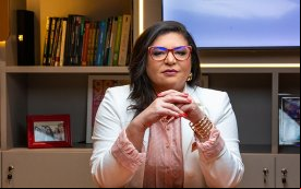 Neiva Ribeiro, presidenta do Sindicato dos Bancários, veste um blazer branco e usa óculos. Está sentada, com os braços apoiados em uma mesa, e dedos das mãos entrelaçados, na altura do peito