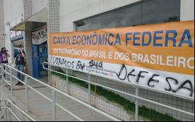 Fachada da agência da Caixa Federal no Parque São Domingos com um faixa do Sindicato estendida, escrita: Caixa Econômoca Federal, um patrimônio do Brasil e dos brasileiros. Na sua parte inferior, a faixa está pixada com a frase: Lutamos em sua defesa. Mais ao fundo da foto, em frente a sua porta, o dirigente André Sardão.  