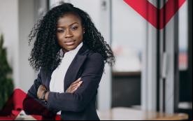 Imagem mostra uma mulher jovem negra, de cabelos compridos e soltos. Ela está com os braços cruzados e usa um terno cinza e uma blusa branca